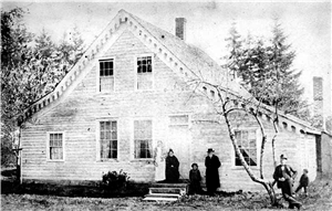 A two story wooden house with decorative roof trim. Four adults stand in front of the house. A child stands off to the right. Another person leans against a tree in the foreground. 